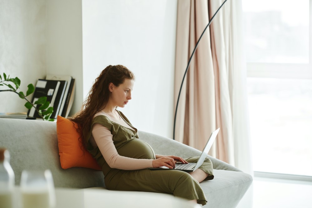 a pregnant women works from home on her couch on a laptop