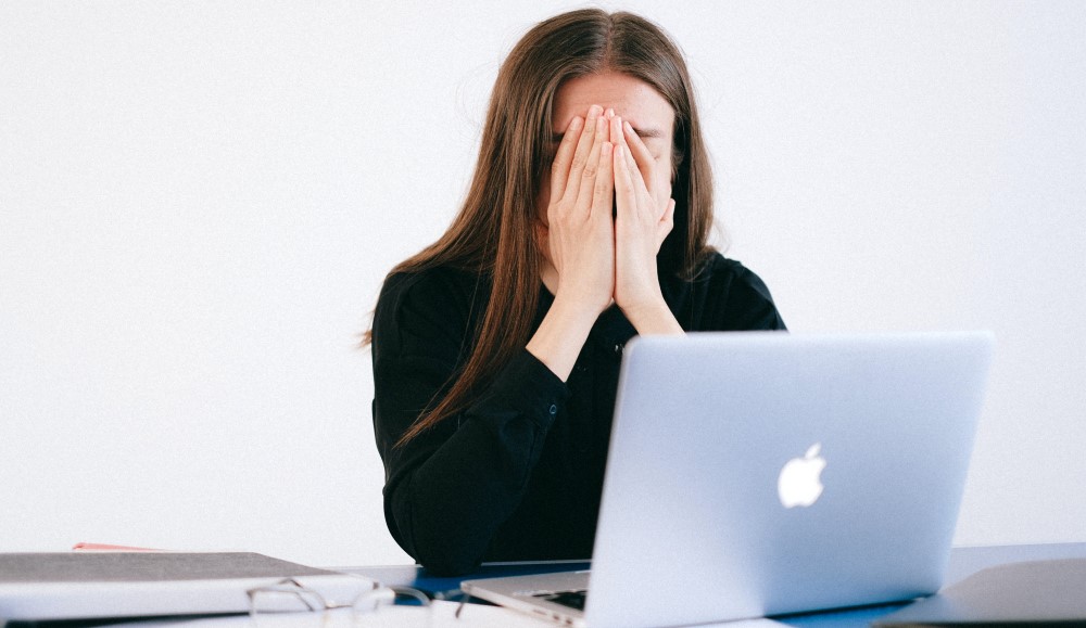 a women puts her hands in her face stressed with a laptop and paper work on a desk